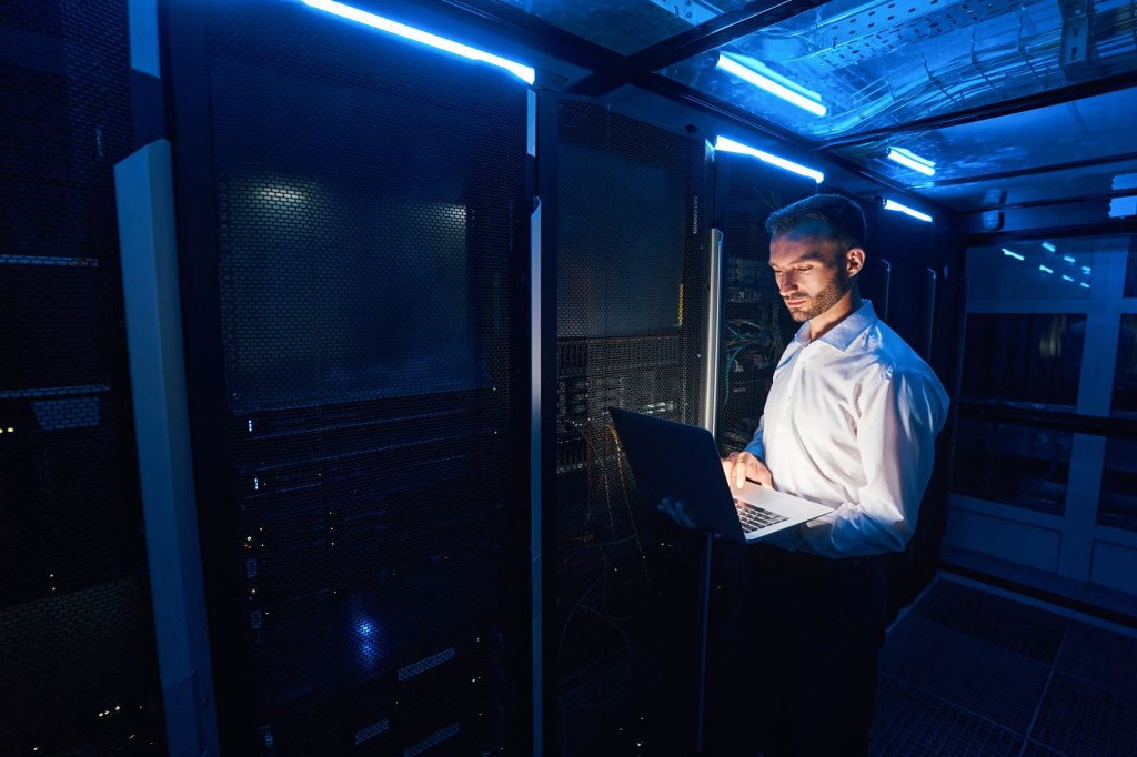 Caucasian man in data center standing near server racks with network equipment and using laptop